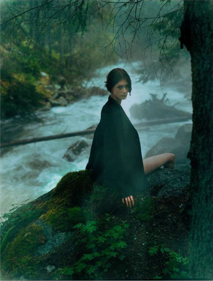 A woman in a dark cloak sits on a mossy rock in a misty forest stream, with a serene gaze and soft, diffused lighting.