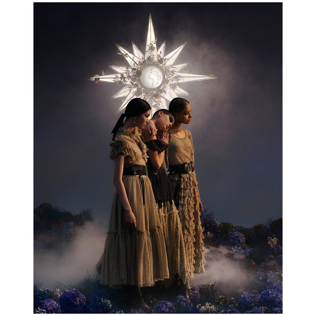 Three fashion models in elegant layered dresses pose under a glowing star-shaped light in a dreamy outdoor setting with flowers and fog.