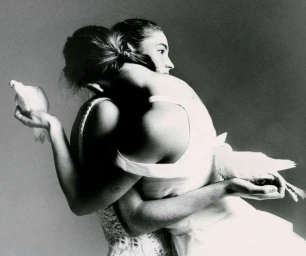 Black and white portrait of two women embracing each other while holding white doves in their outstretched hands against a grey background.