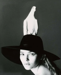 Portrait of a woman wearing a wide black brimmed hat with a white dove perched on top, photographed in monochrome.