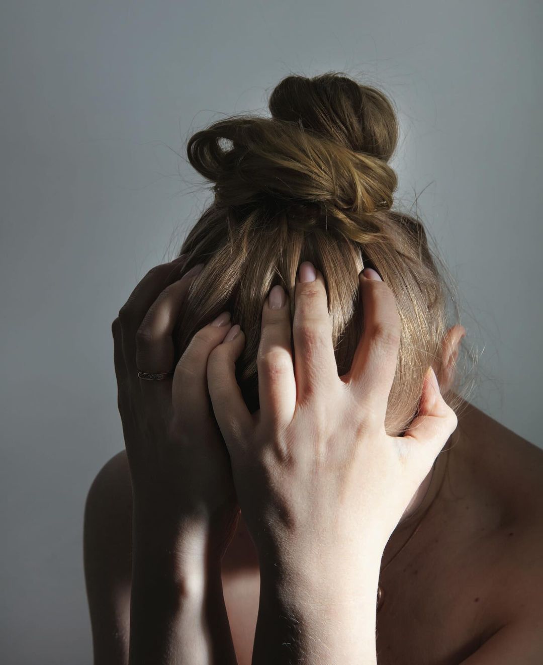 Close-up studio portrait of a fair-skinned adult female with auburn hair in a messy bun, her hands covering her face against a gray backdrop.