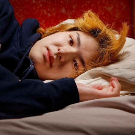 Close-up portrait of an adult with orange-blonde hair lying down indoors, wearing a dark hoodie, looking directly at the camera. 