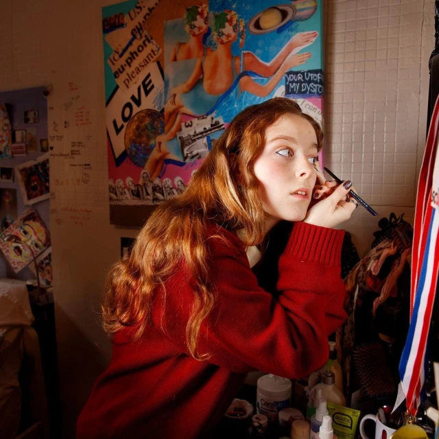 Young adult female applying makeup in a cluttered room with a vibrant art poster and various cosmetic items, indoors.