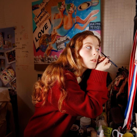 Young adult female applying makeup in a cluttered room with a vibrant art poster and various cosmetic items, indoors.