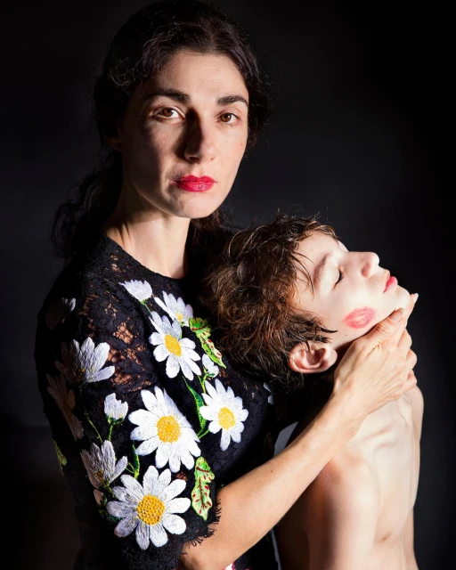 A woman in a daisy sweater embraces a child with red lipstick kiss marks on their face in a dark studio portrait.
