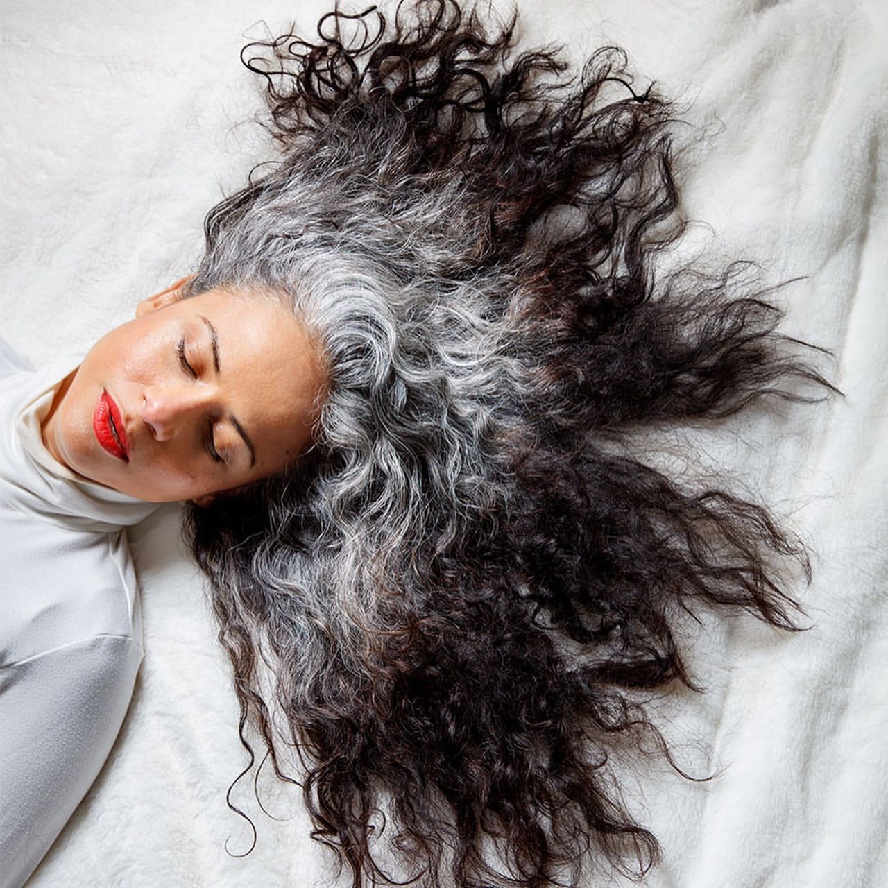 Overhead shot of a woman with curly black and grey hair lying on a white, textured surface with red lipstick and a white turtleneck.