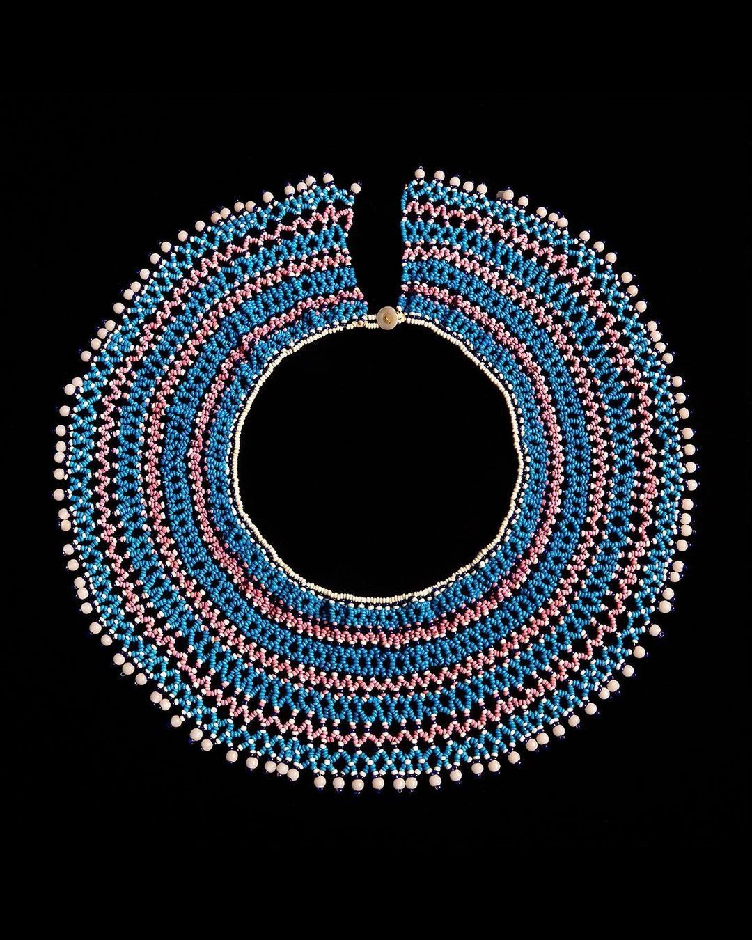 Studio shot of a colorful beaded collar necklace with pink, blue, and white beads arranged in a pattern on a dark black background.