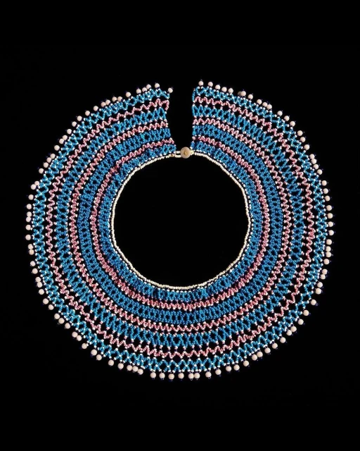 Studio shot of a colorful beaded collar necklace with pink, blue, and white beads arranged in a pattern on a dark black background.