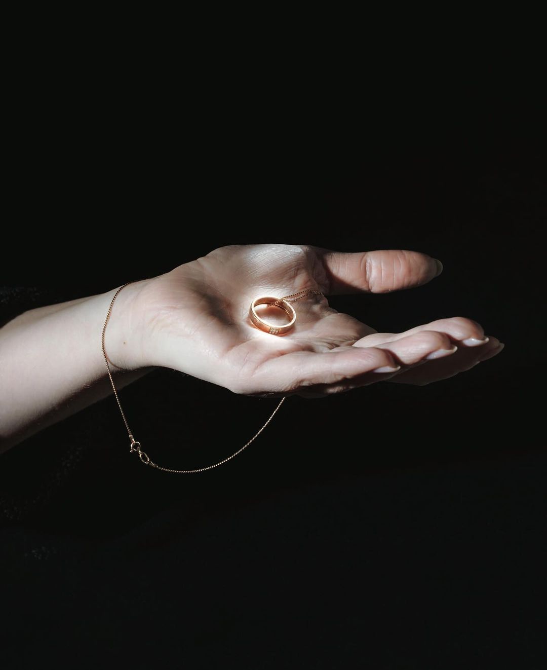 Hand holding gold ring with pendant necklace against black background; high contrast lighting accentuates the metallic jewelry.