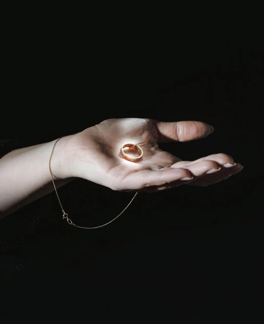 Hand holding gold ring with pendant necklace against black background; high contrast lighting accentuates the metallic jewelry.