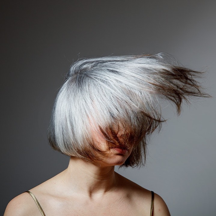Portrait of a person with grey and brown hair blowing away from their face, against a neutral grey background in a studio setting.