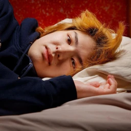 Close-up portrait of a person with dyed orange hair lying in bed wearing a navy blue hoodie, photographed by Elinor Carucci.