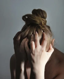 Close-up portrait of a woman holding her head in her hands with brown hair in a messy bun against a grey background