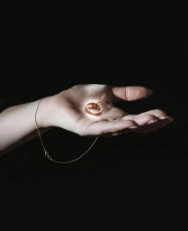 Close-up of a person's open palm holding a glowing gold ring with a thin gold chain resting against the wrist on a dark background.