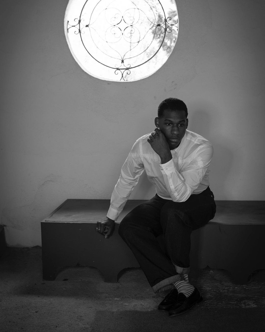 Monochrome portrait of a Black man sitting on a bench with a contemplative expression, indoors, illuminated by light from a round window.