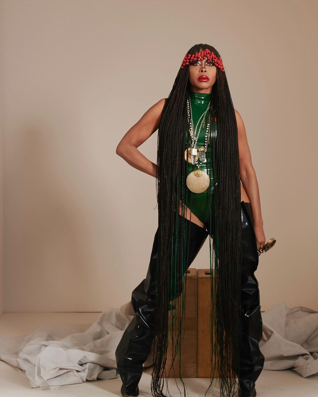 Woman with beaded headpiece poses in a green latex bodysuit with silver necklaces, black boots, and long dark braids in a studio.
