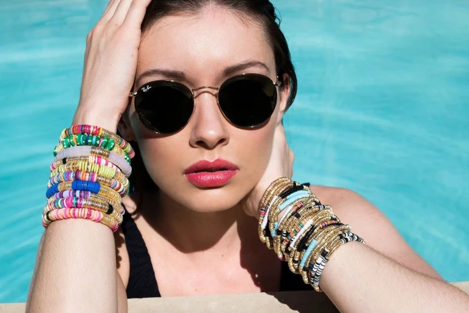 Woman wearing round sunglasses and colourful stacked bracelets by a blue pool, photographed by Elizabeth Lavin.