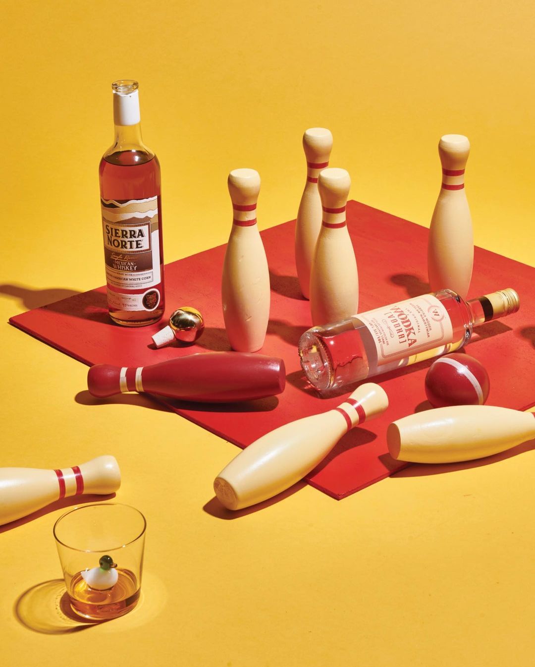 Still life composition with vodka and whiskey bottles, a glass, and bowling pins arranged on a yellow background with a red surface.