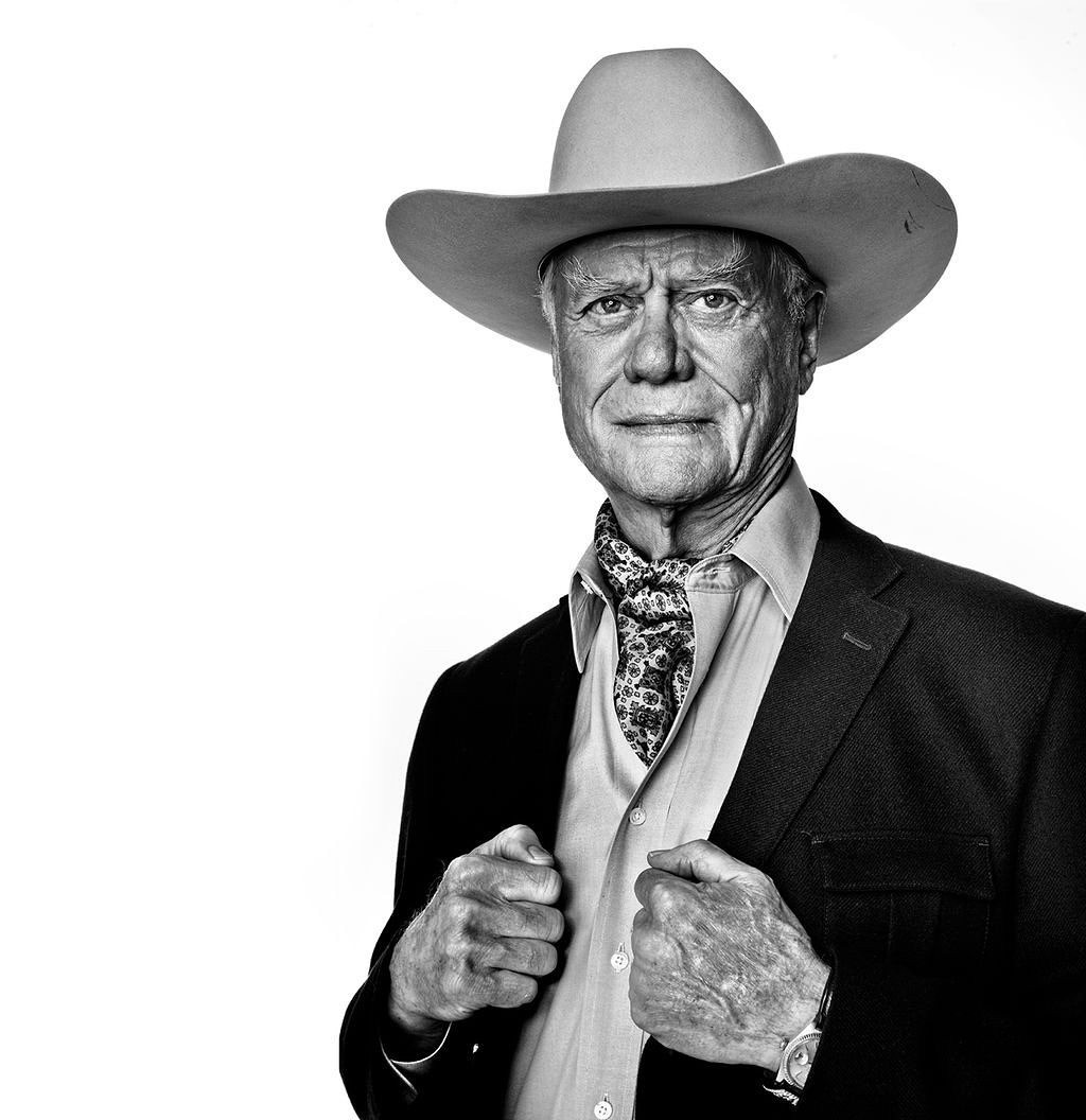 Portrait of an elderly man wearing a cowboy hat, patterned neckscarf, and a suit jacket against a white background.