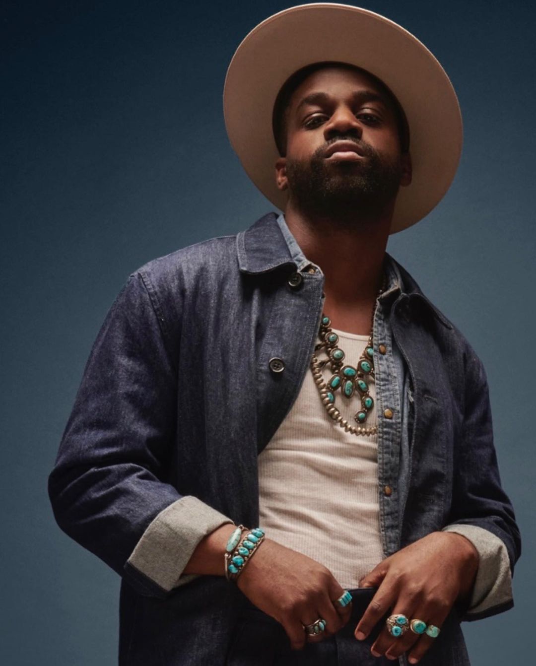 Man wearing a hat and denim jacket poses in a medium shot with turquoise jewellery against a blue studio backdrop, fashion portrait.