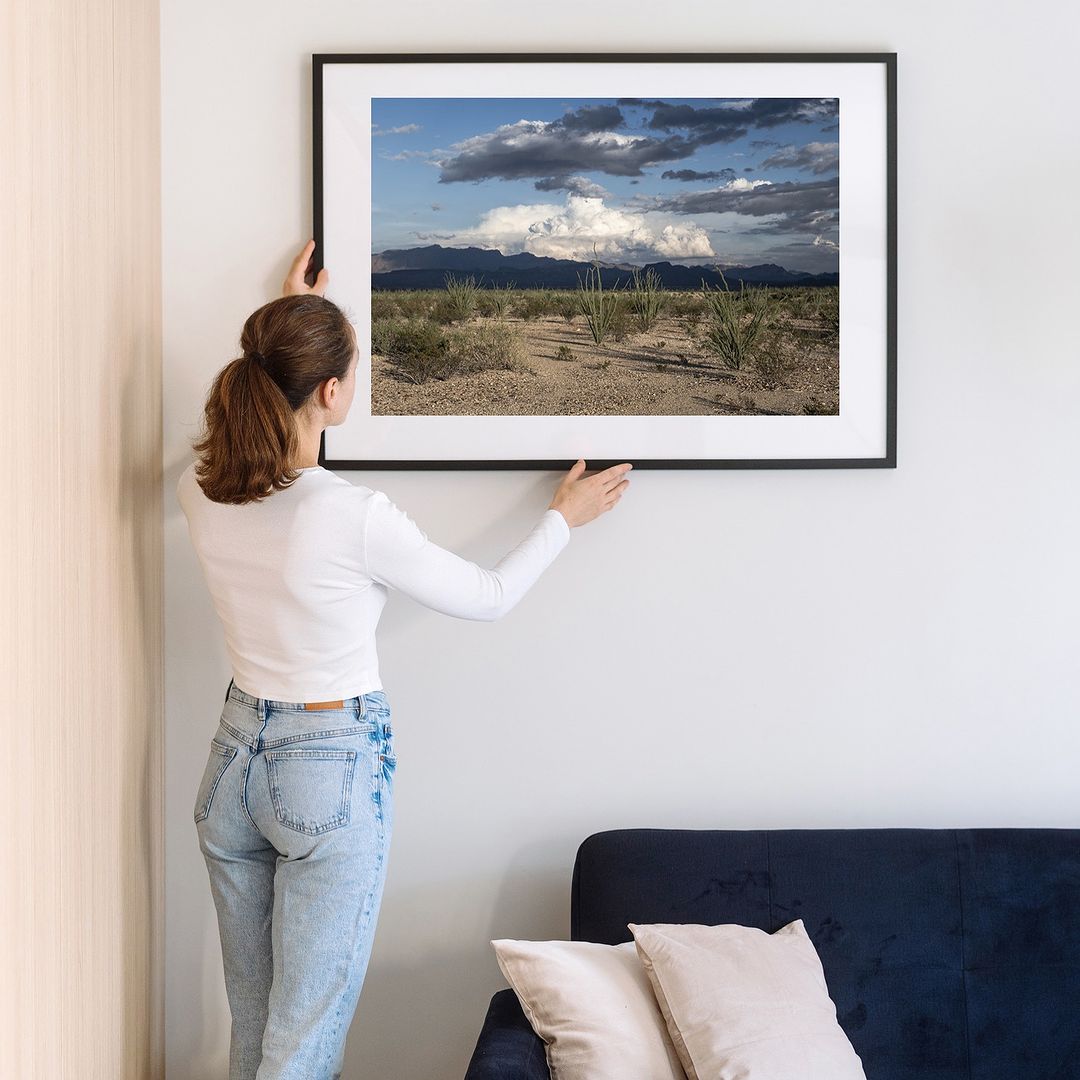 Framed desert landscape art print on a wall, woman adjusts the frame above a navy couch with beige pillows, minimalist interior design.