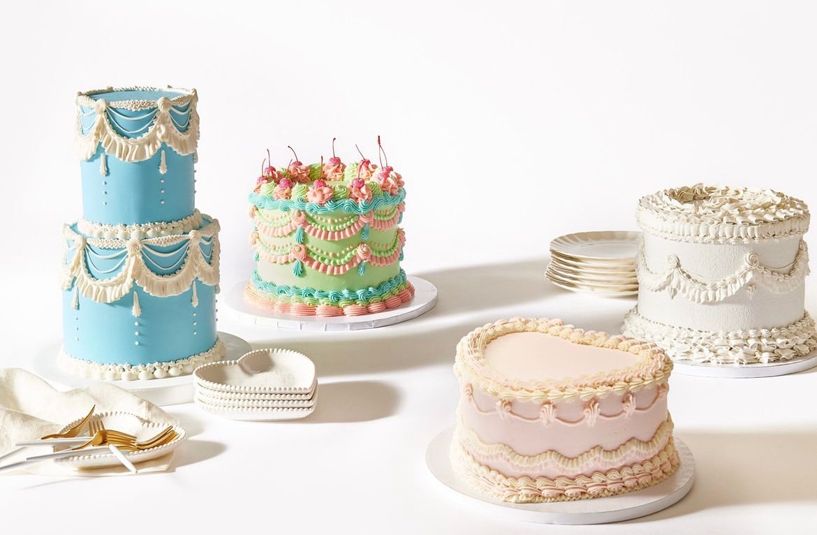 Four decorative cakes with colorful icing in a variety of shapes and sizes on a white surface, with small plates and dessert forks.