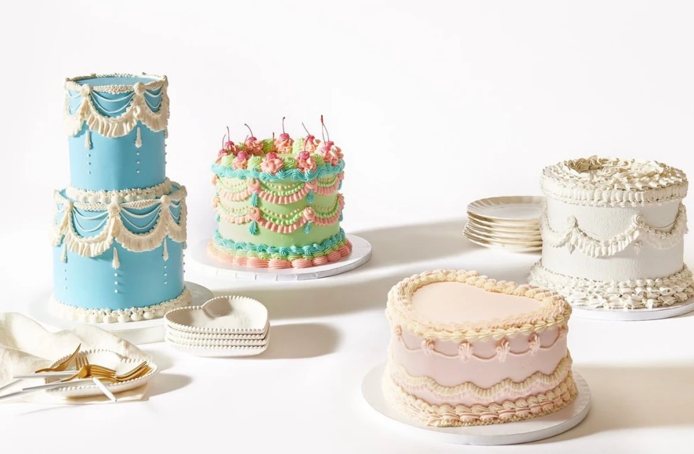 Four decorative cakes with colorful icing in a variety of shapes and sizes on a white surface, with small plates and dessert forks.