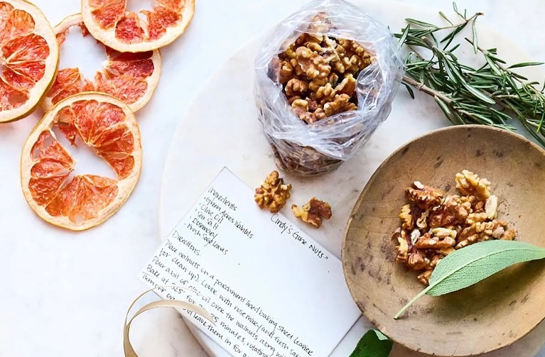 Overhead shot of glazed walnuts, grapefruit slices, rosemary, and a recipe card, set on a white surface with a wooden bowl and sage leaf.