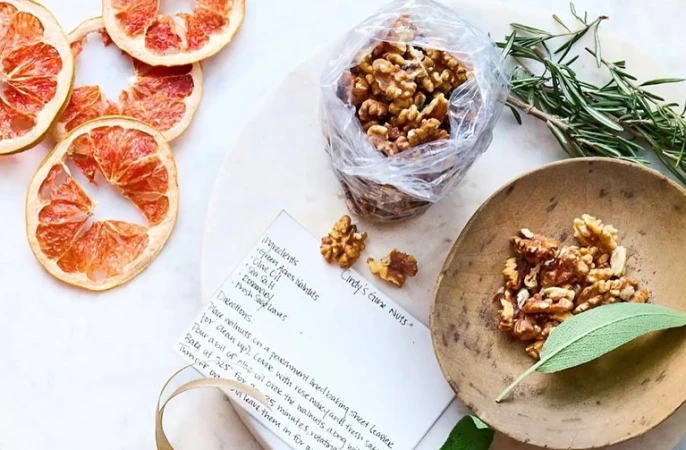 Overhead shot of glazed walnuts, grapefruit slices, rosemary, and a recipe card, set on a white surface with a wooden bowl and sage leaf.
