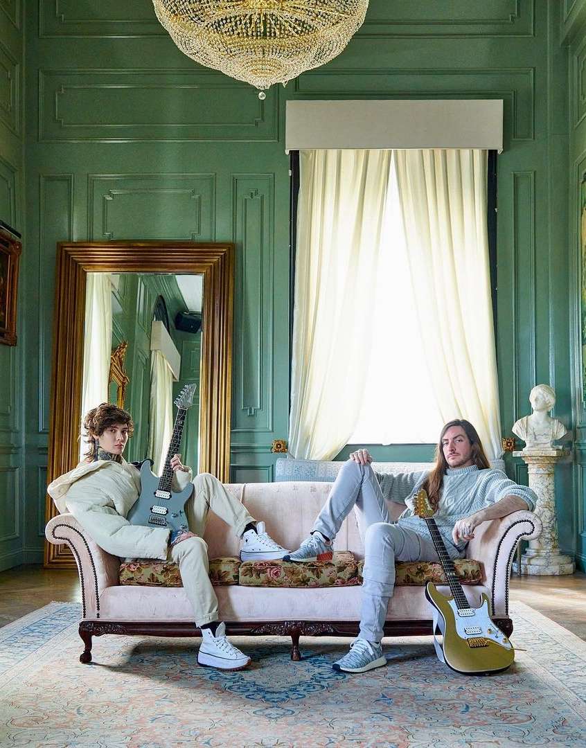 Two adult males pose with electric guitars on a floral couch inside a room with green paneled walls, a chandelier, and a rug.