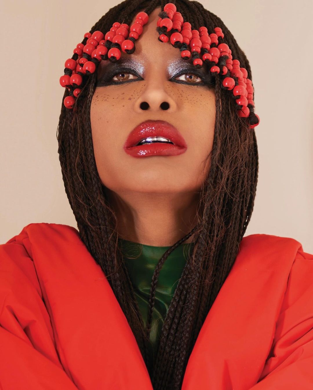 Woman with red beads in braids, wearing glossy red lipstick and a puffy red jacket, looks upward in a studio portrait.