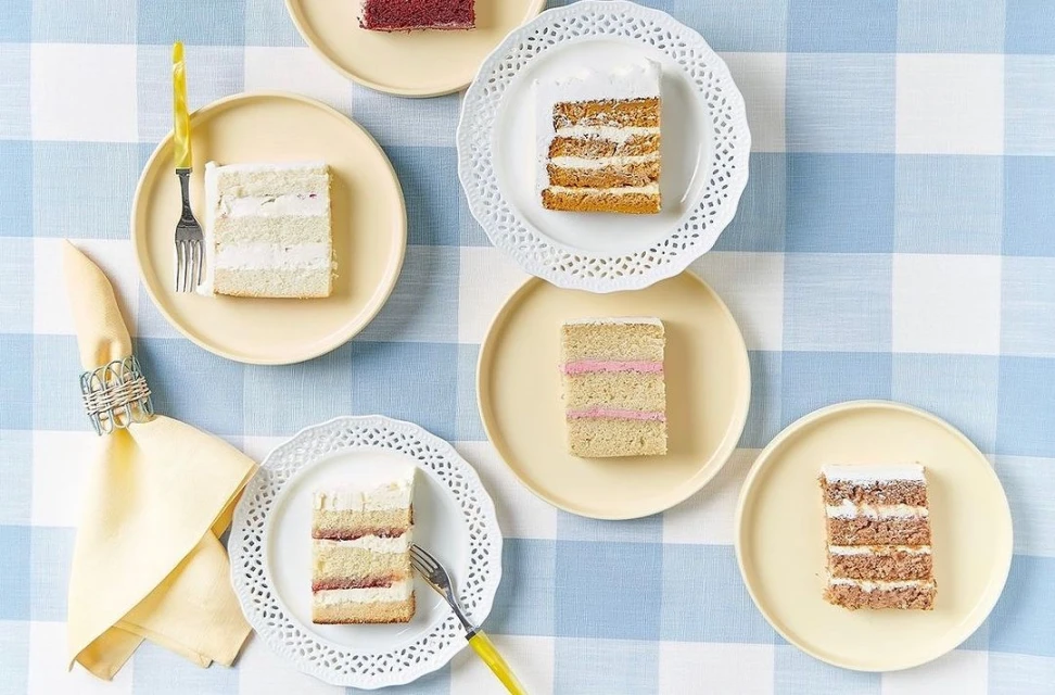 Overhead shot of multiple cake slices on plates with silverware, arranged on a blue and white checkered tablecloth.