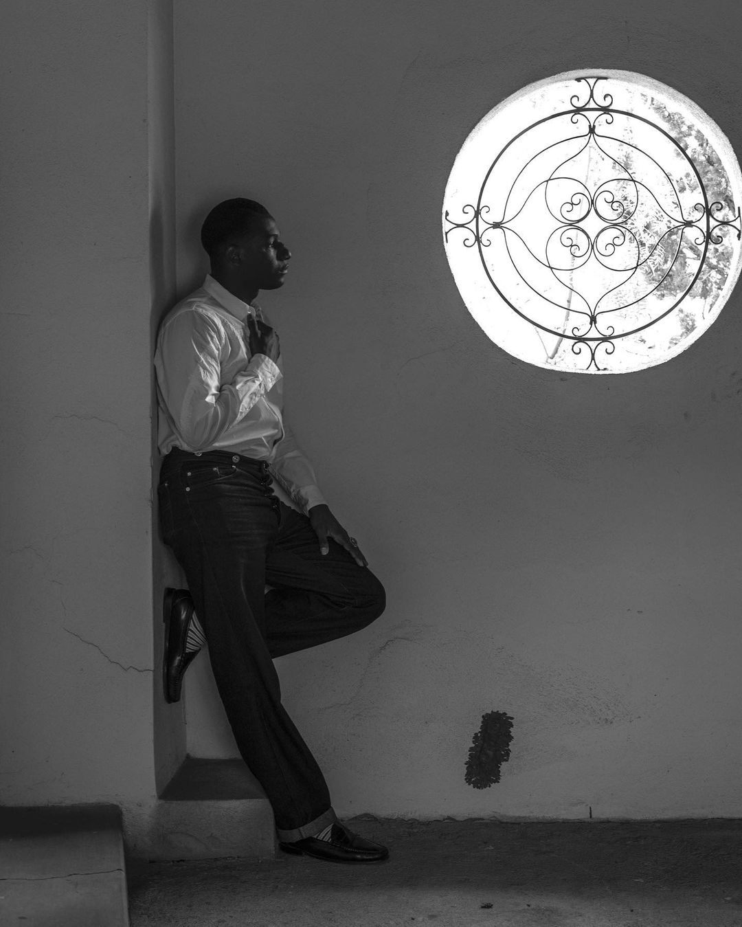 Portrait of a Black man in a white shirt leaning against a wall in front of a round window with decorative metalwork in black and white.