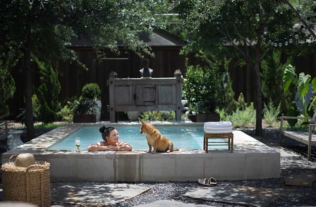 Woman relaxing in a backyard pool with a dog, enjoying a drink on a sunny day in a serene outdoor setting.