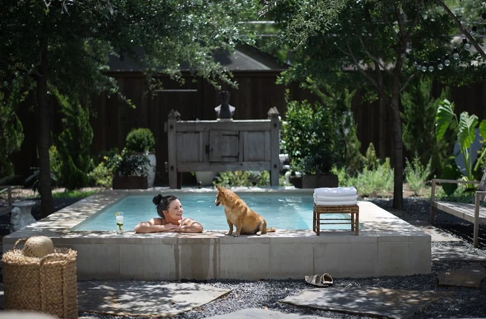 Woman relaxing in a backyard pool with a dog, enjoying a drink on a sunny day in a serene outdoor setting.