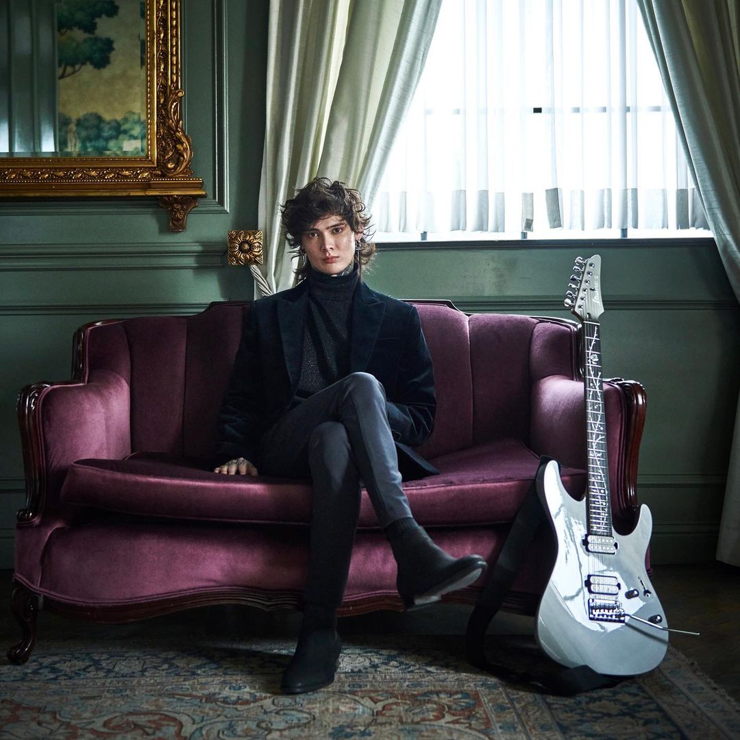 Stylish man poses on a plush purple couch indoors with a silver electric guitar in a room with dark green walls and curtains.