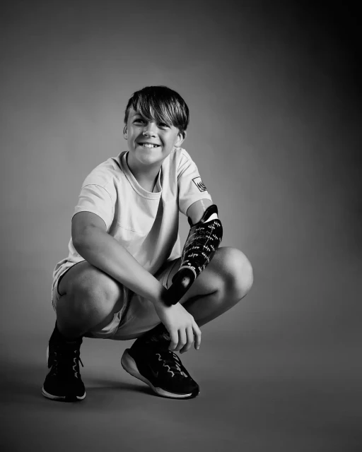 Portrait of a young boy with prosthetic arm squatting in a studio, looking up and smiling against a graduated grey background.