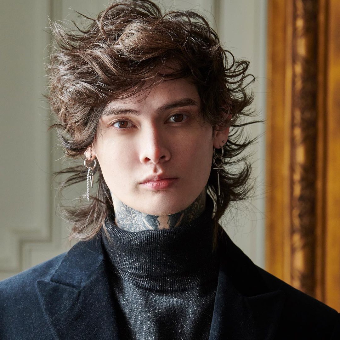 Close-up portrait of an adult with wavy brown hair, wearing a turtleneck sweater and a blazer, accented with silver earrings and neck tattoos.