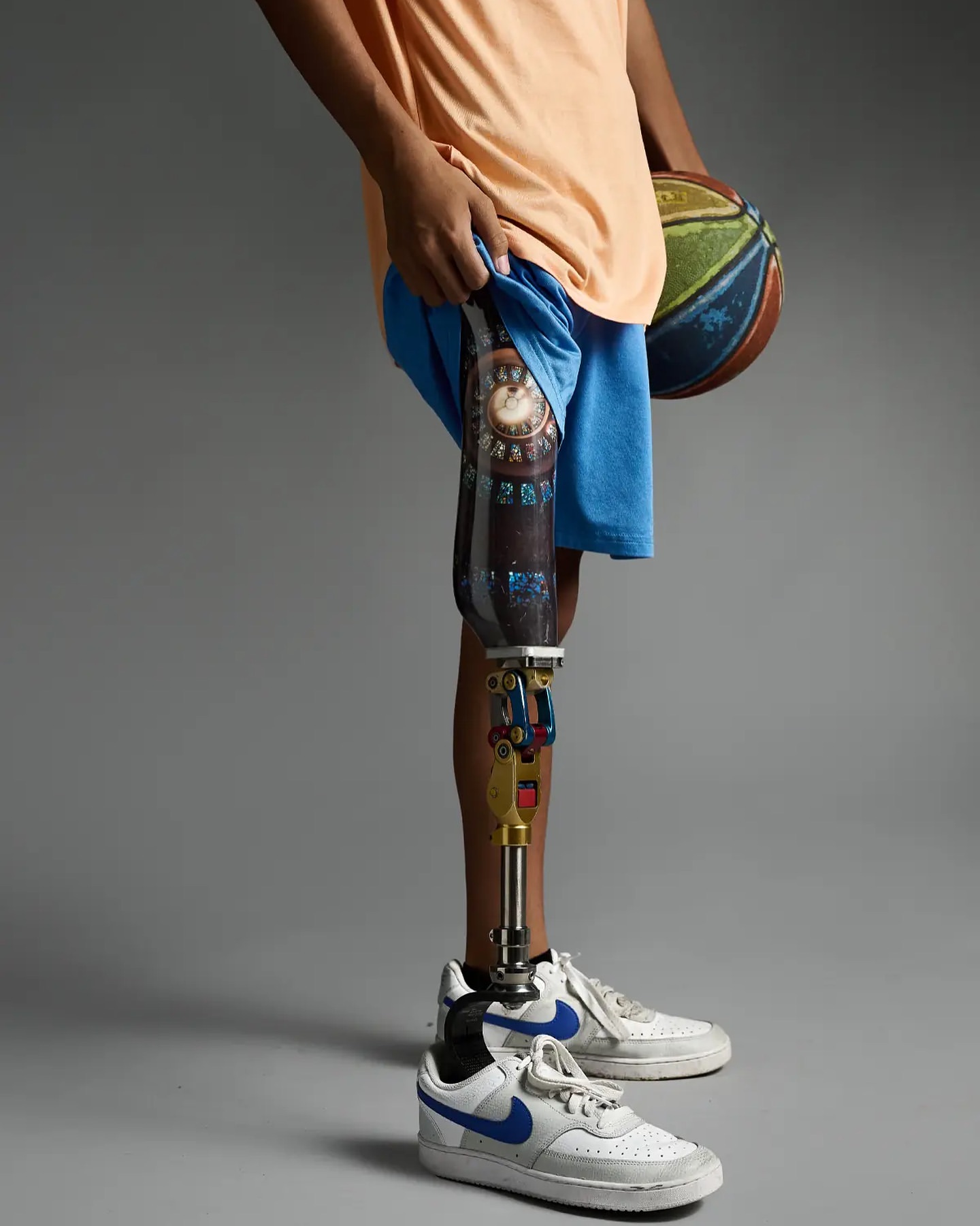 Young man stands in a studio with a basketball and a decorative prosthetic leg, wearing athletic shorts and sneakers.