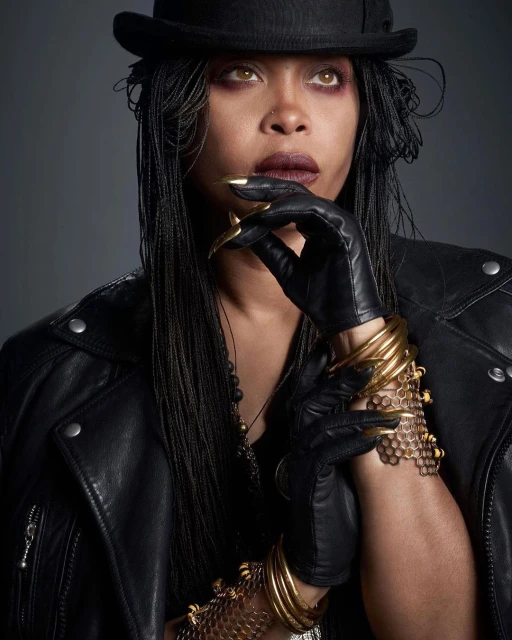 Portrait of a woman with braided hair, wearing a black hat and jacket, adorned with gold bracelets, against a grey backdrop.