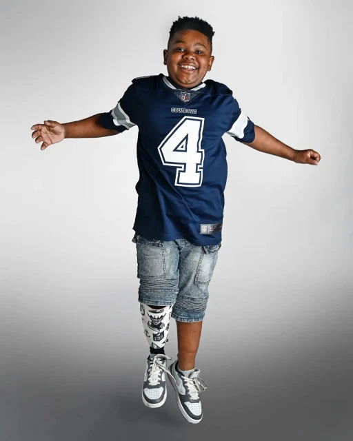 Young boy in Dallas Cowboys jersey leaps in photo studio with a prosthetic leg, smiling and full of energy against a gradient background.