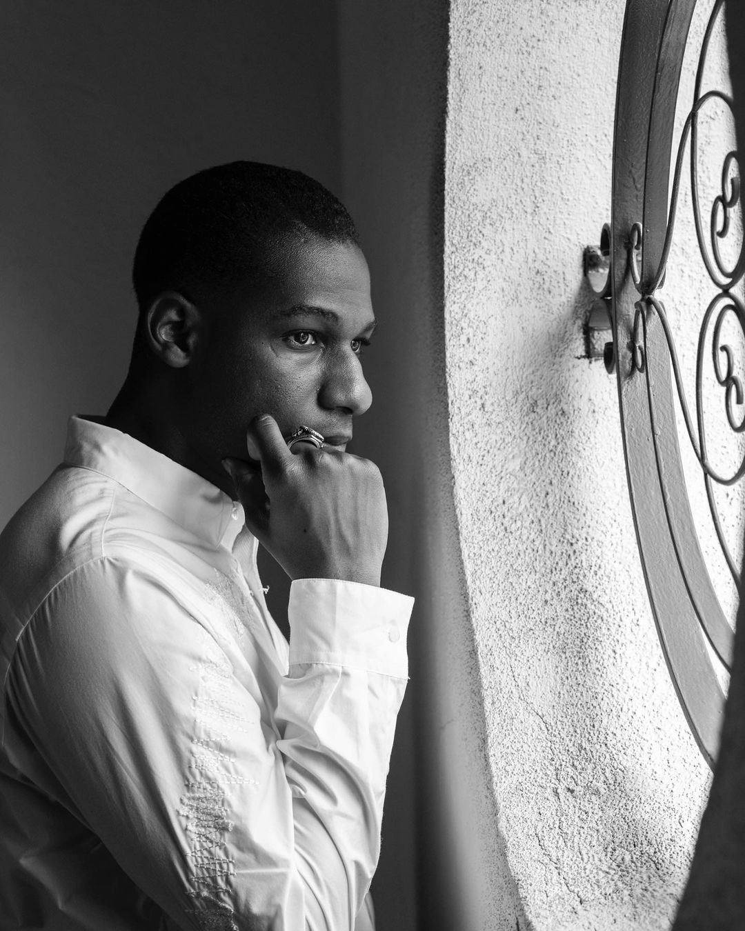 Monochrome portrait of a Black adult male in a white collared shirt with hand on chin, gazing thoughtfully near a decorative window.