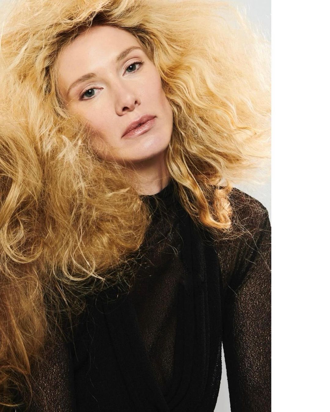 Portrait of a blonde adult woman with dramatic wavy hair wearing a semi-sheer top against a white background in an indoor setting.