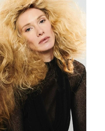 Portrait of a blonde adult woman with dramatic wavy hair wearing a semi-sheer top against a white background in an indoor setting.