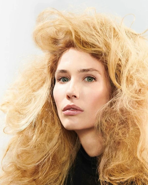 Beauty shot of a blond-haired woman in a black top against a bright white backdrop. The woman has a fair complexion and light green eyes.