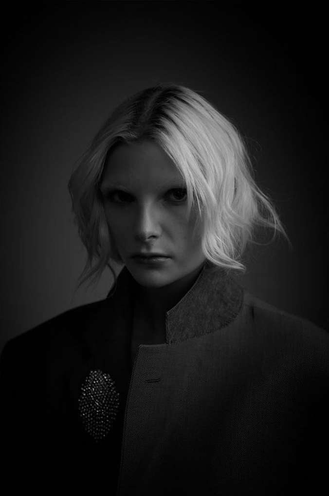 Monochrome portrait of an adult woman with blonde hair wearing a jacket with an embellished brooch, captured in a photo studio.