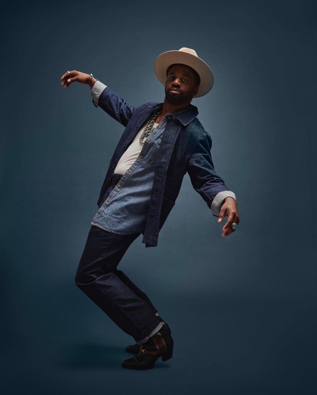 A Black male model poses indoors wearing a denim jacket, cream-colored hat, jeans and cowboy boots against a dark backdrop in a studio shot.