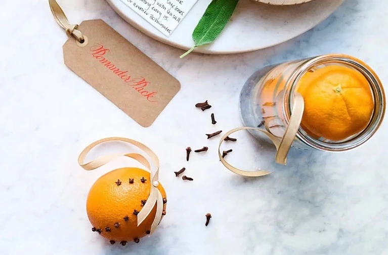 Overhead shot of homemade orange pomander balls with cloves, ribbon, and a craft tag, arranged on a white marble surface.