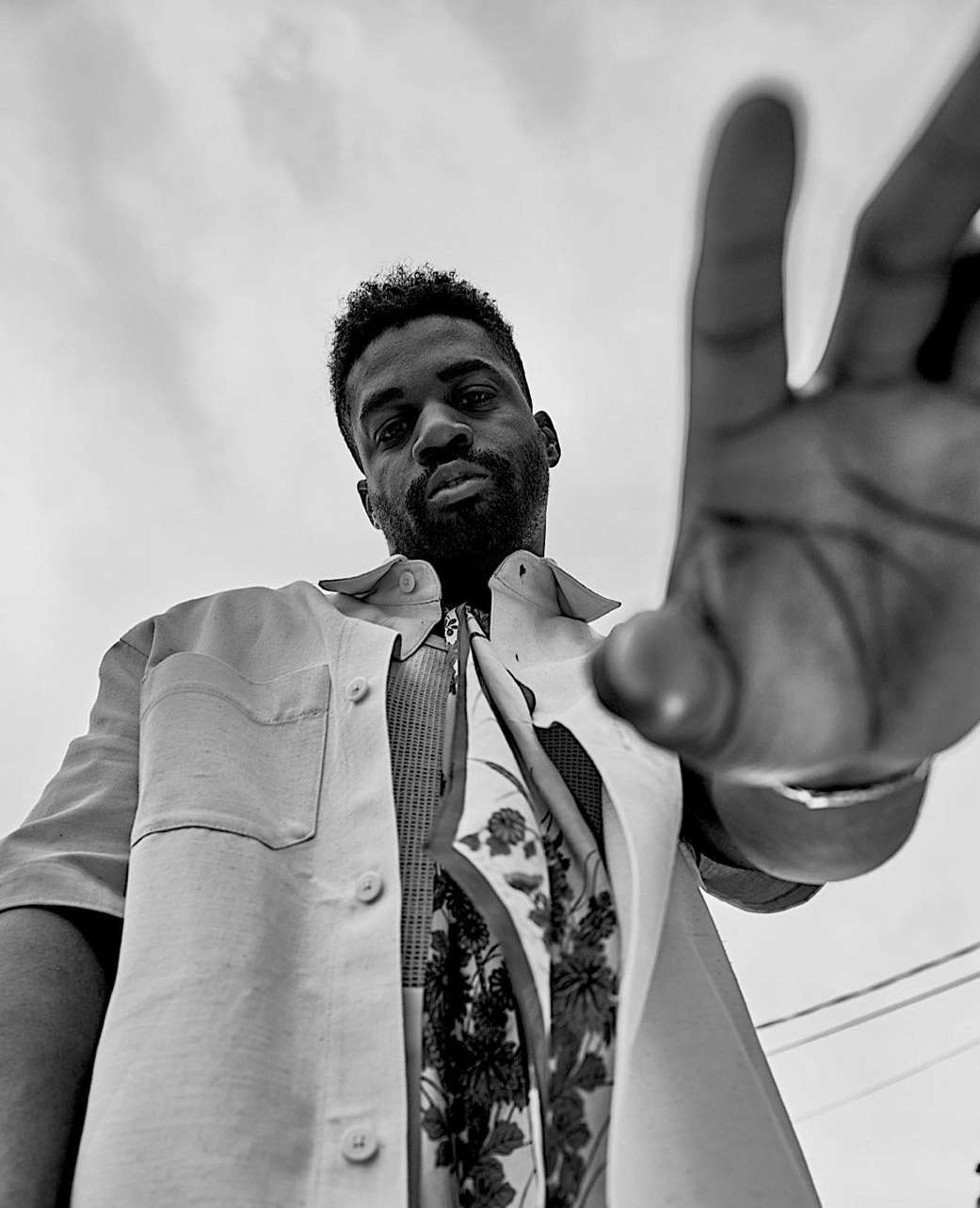 Striking black-and-white portrait of an African American man looking down with hand raised, wearing an unbuttoned casual shirt and patterned top.