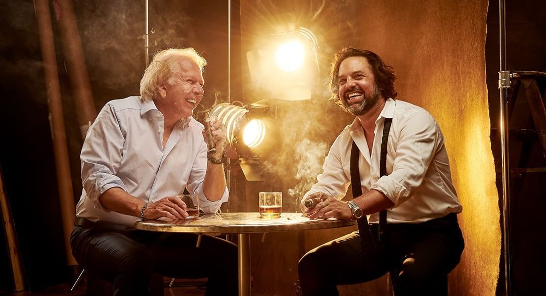 Two men sitting at a table with drinks and cigars in a studio setting with warm lighting and smoke effects for a sophisticated portrait.
